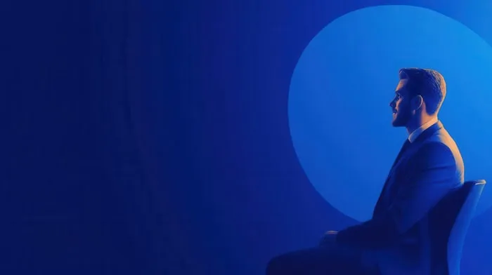 Hombre con traje sentado de perfil sobre un fondo azul con un círculo luminoso detrás