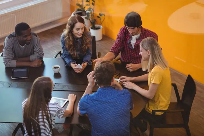 Seis personas reunidas alrededor de una mesa de oficina con tabletas y tazas de café, ante una pared naranja